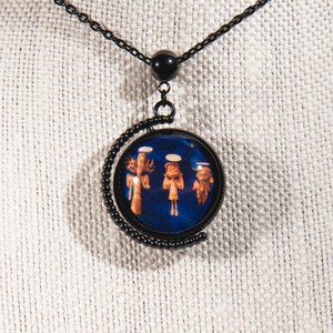 Puede incluir: Un collar de cadena negra con un colgante redondo que presenta un fondo azul con tres &aacute;ngeles con t&uacute;nicas blancas. El colgante est&aacute; enmarcado por una luna creciente de metal negro.
