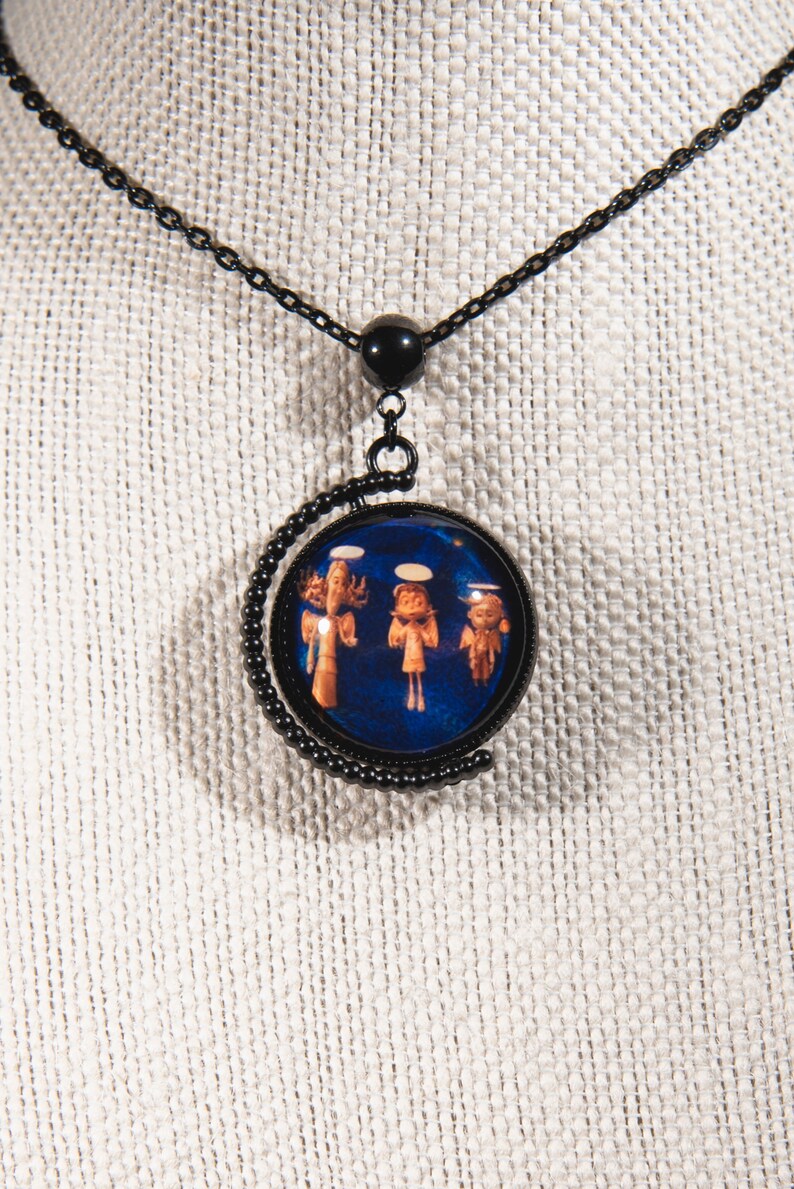 Puede incluir: Un collar de cadena negra con un colgante redondo que presenta un fondo azul con tres &aacute;ngeles con t&uacute;nicas blancas. El colgante est&aacute; enmarcado por una luna creciente de metal negro.