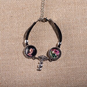 May include: Black leather bracelet with two round silver charms featuring colorful floral designs. The charms are connected by a silver chain with a small silver charm.
