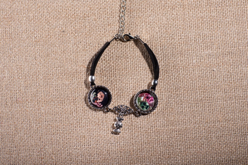 May include: Black leather bracelet with two round silver charms featuring colorful floral designs. The charms are connected by a silver chain with a small silver charm.