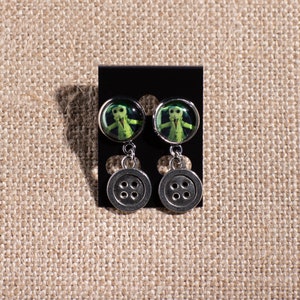 May include: A pair of silver dangle earrings with a green button-shaped charm featuring a character from the movie "The Nightmare Before Christmas".