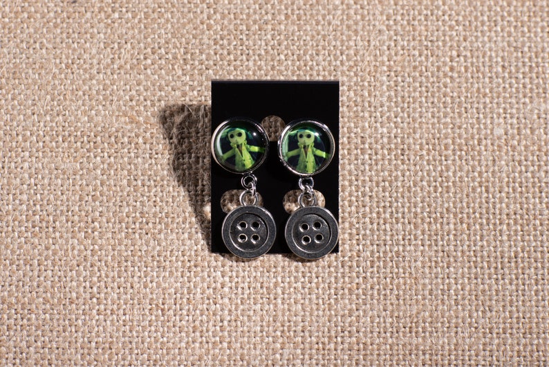 May include: A pair of silver dangle earrings with a green button-shaped charm featuring a character from the movie "The Nightmare Before Christmas".