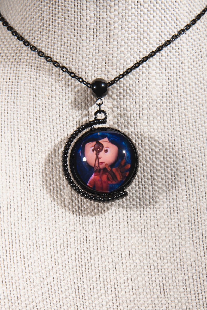 Puede incluir: Collar negro con un colgante redondo de vidrio que presenta un personaje de la pel&iacute;cula "Coraline". El personaje es una ni&ntilde;a con cabello azul y un vestido rojo, sosteniendo una llave.