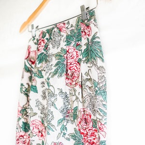May include: A white skirt with a floral print featuring red, pink, and green flowers. The skirt is made of a lightweight fabric and has a pleated design.