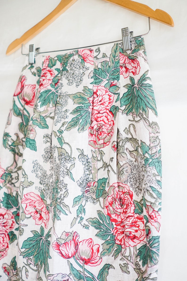 May include: A white skirt with a floral print featuring pink and green flowers. The skirt has a pleated design and is made of a lightweight fabric.