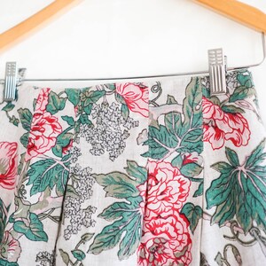 May include: A white skirt with a floral print featuring pink, red, and green flowers. The skirt has pleats and is hanging on a wooden hanger.