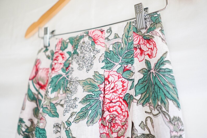 May include: A white linen skirt with a floral print featuring pink and green flowers. The skirt has pleats and is hanging on a wooden hanger.