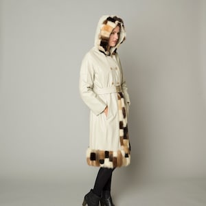 Puede incluir: Abrigo con capucha color crema con cinturón y ribete de piel. El abrigo presenta un diseño cruzado con botones dorados. La capucha y el dobladillo están adornados con un patchwork de piel marrón, beige y crema. El abrigo se lleva con pantalones y botas negras.