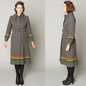 Puede incluir: Un abrigo largo gris con cinturón y rayas de colores en los puños y el dobladillo. El abrigo tiene cuello y cierre de botones.