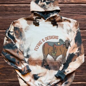 May include: A hooded sweatshirt with a bleached, tie-dye effect in shades of brown, black, and cream. The front features a graphic of a bull with a desert landscape and the text "FLYING'S DESIGNS". The cuffs and hood lining are dark gray.