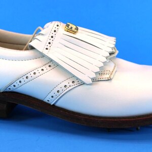 May include: A white leather golf shoe featuring a decorative kiltie fringe over the laces, a gold-toned metal emblem on the fringe, brogue detailing along the side panels, and a dark brown stacked heel with a textured sole for traction.