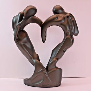 May include: A dark brown, matte-finished sculpture depicting two abstract human figures leaning toward each other to form a heart shape. The figures are connected at the base and at the top, creating a continuous, fluid silhouette with smooth curves.