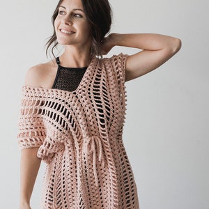 Puede incluir: Una prenda de vestir de crochet rosa con un lazo en la cintura y aberturas laterales. La prenda está hecha con un delicado patrón de punto abierto.