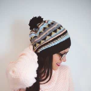 May include: A black, gray, and light blue patterned knit beanie with a black pom-pom. The beanie is worn with a light pink sweater. The text "MEGMADE with love" is visible in white.