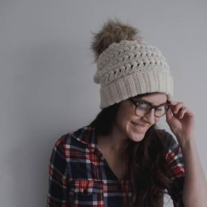 May include: A woman wearing a white crocheted beanie with a faux fur pom pom and a red and blue plaid shirt. She is smiling and holding her glasses in her hand.