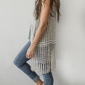 Pode incluir: Um colete de crochê bege com trama aberta e detalhes de franjas. O colete comprido é sobreposto a um top azul claro e combinado com jeans azuis. A modelo está em pé contra uma parede branca.