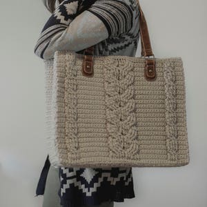 Puede incluir: Un bolso tote de crochet beige con asas de cuero marrón. El bolso tiene un patrón de punto de cable y es lo suficientemente grande como para llevar los artículos esenciales del día a día.
