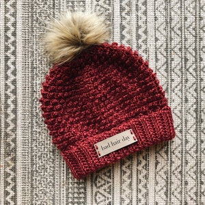 May include: A red crocheted beanie with a brown faux fur pom pom. The beanie has a brown leather patch with the text "bad hair day" on it.