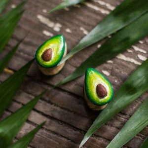 May include: Two avocado-shaped wooden earrings with green and brown paint. The earrings are on a wooden surface with green leaves.