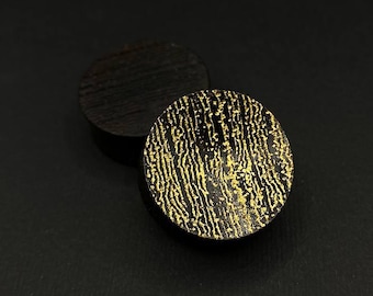 Op de afbeelding: Twee donkerbruine, ronde houten pluggen met een goudkleurig, getextureerd ontwerp op de voorkant. Eén plug is scherp, terwijl de andere enigszins onscherp is op de achtergrond. De pluggen staan tegen een zwarte achtergrond.