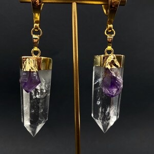 May include: Gold-coloured earrings featuring clear crystal pendants with raw, purple amethyst accents. The earrings are suspended from gold hooks and displayed on a gold stand against a dark backdrop.