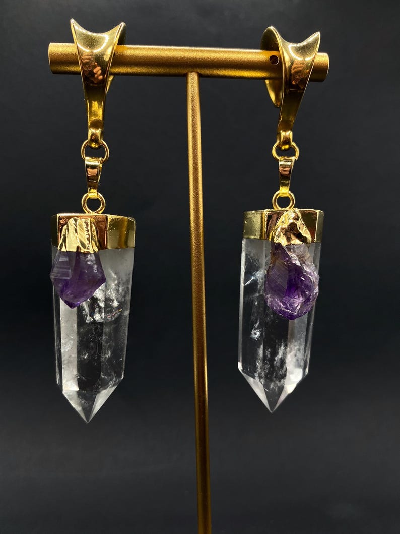 May include: Gold-coloured earrings featuring clear crystal pendants with raw, purple amethyst accents. The earrings are suspended from gold hooks and displayed on a gold stand against a dark backdrop.