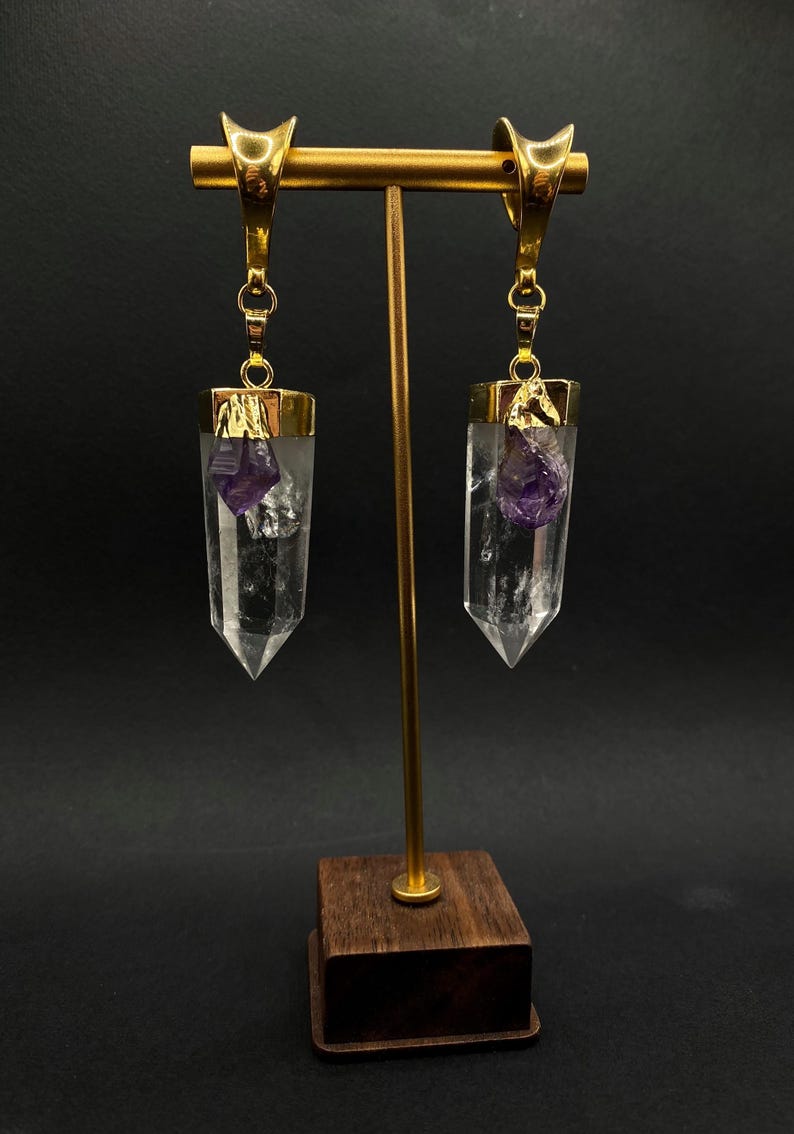 May include: Gold-coloured earrings featuring clear quartz crystal pendants, each with a small, purple amethyst crystal. The earrings are displayed on a gold stand with a wooden base. The quartz crystals are approximately 5 cm long.