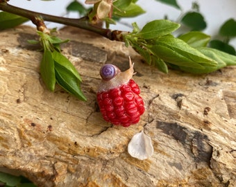 May include: A small snail with a purple shell is perched on top of a red raspberry. The raspberry is sitting on a piece of bark with a white petal nearby.