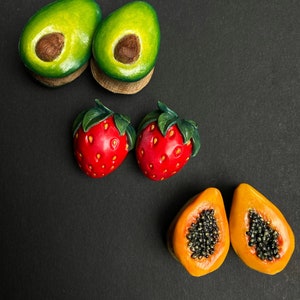 May include: A set of six fruit-shaped wooden earrings. The earrings are in the shape of avocados, strawberries, and papayas. The avocados are green with brown pits, the strawberries are red with green leaves, and the papayas are orange with black seeds.