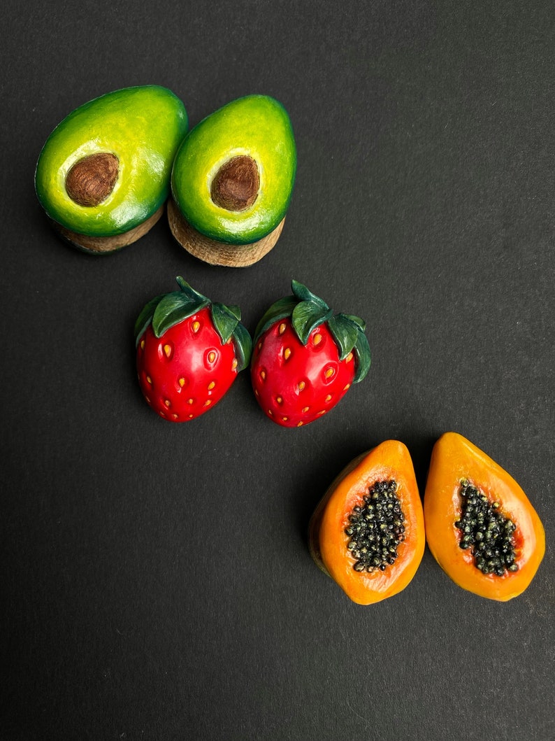 May include: A set of six fruit-shaped wooden earrings. The earrings are in the shape of avocados, strawberries, and papayas. The avocados are green with brown pits, the strawberries are red with green leaves, and the papayas are orange with black seeds.