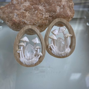 May include: Two wooden earrings featuring a carved design of white mushrooms and a clear quartz crystal. The earrings are oval shaped and have a natural wood finish.
