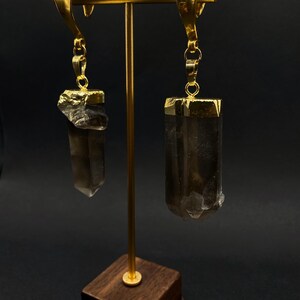 May include: Gold earrings with smoky quartz crystals. Each earring has a gold-plated cap on the crystal, suspended from a decorative gold hook. The crystals are a translucent brown colour, displayed on a gold stand with a wooden base.