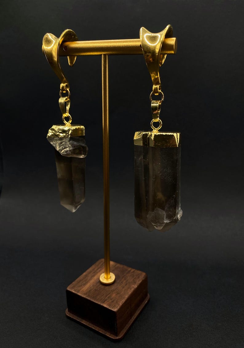 May include: Gold earrings with smoky quartz crystals. Each earring has a gold-plated cap on the crystal, suspended from a decorative gold hook. The crystals are a translucent brown colour, displayed on a gold stand with a wooden base.