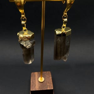May include: Gold-coloured earrings with a distinctive design. Each earring has a gold-plated top and a smoky quartz crystal pendant. The crystals have a rough, natural texture and hang from delicate gold chains. The earrings are displayed on a gold stand with a wooden base.