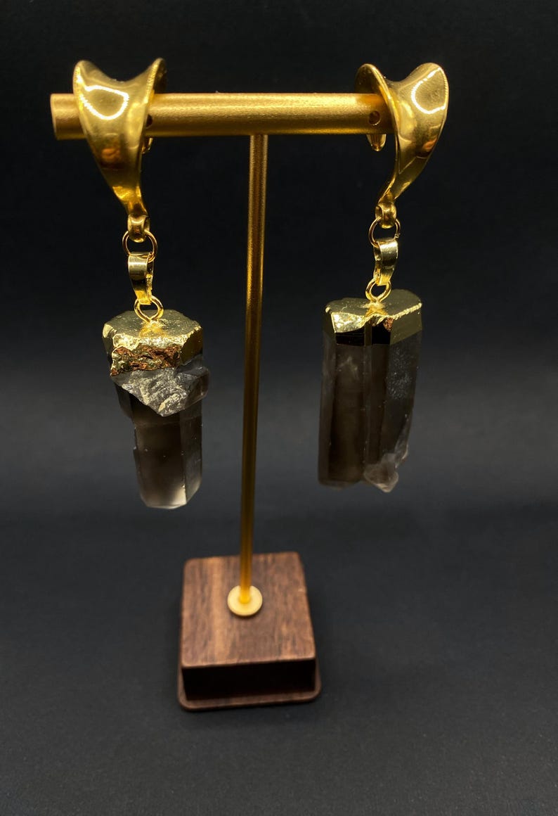 May include: Gold-coloured earrings with a distinctive design. Each earring has a gold-plated top and a smoky quartz crystal pendant. The crystals have a rough, natural texture and hang from delicate gold chains. The earrings are displayed on a gold stand with a wooden base.