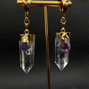 May include: Clear quartz crystal earrings with gold-coloured metal settings and accents. Each earring features a raw amethyst crystal. The earrings are displayed on a gold-coloured stand against a dark background.