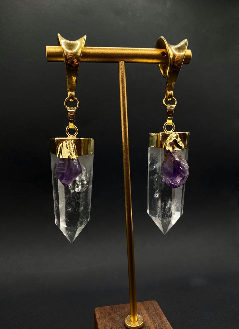 May include: Clear quartz crystal earrings with gold-coloured metal settings and accents. Each earring features a raw amethyst crystal. The earrings are displayed on a gold-coloured stand against a dark background.