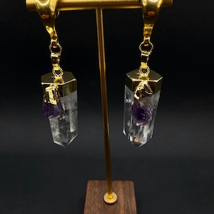 May include: Gold-coloured earrings with clear crystal pendants, each with a hexagonal gold cap and a small purple amethyst stone. The earrings hang from a gold bar on a wooden stand, against a dark background.