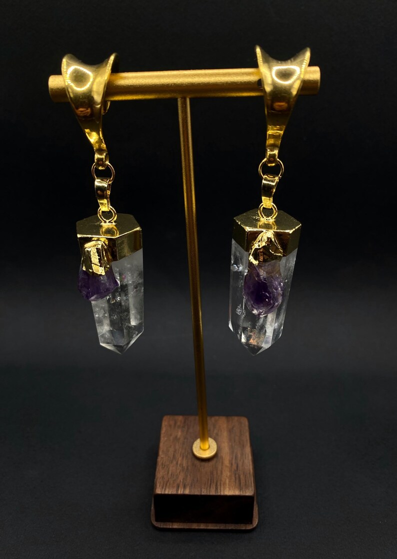 May include: Gold-coloured earrings with clear crystal pendants, each with a hexagonal gold cap and a small purple amethyst stone. The earrings hang from a gold bar on a wooden stand, against a dark background.