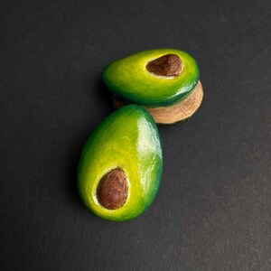 May include: Two avocado halves painted on a dark background. The avocado halves are green with brown pits.