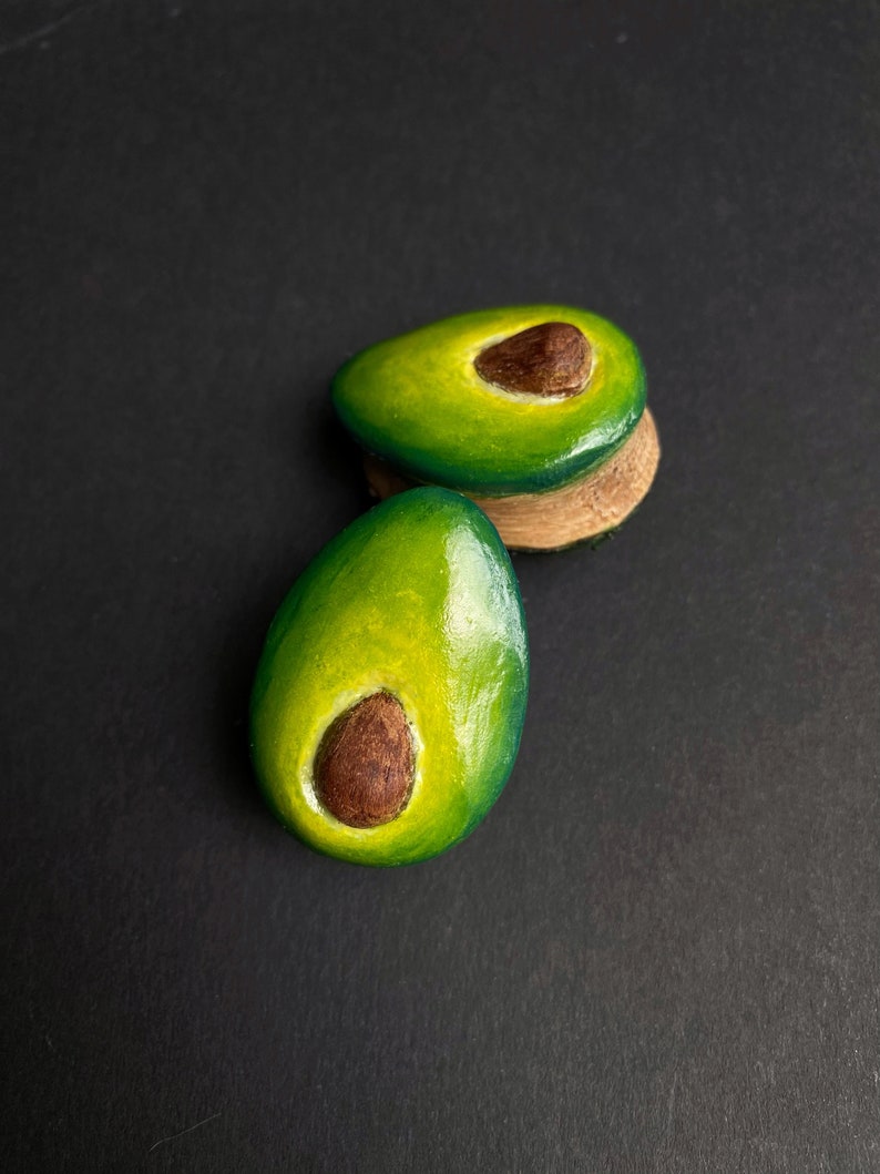 May include: Two avocado halves painted on a dark background. The avocado halves are green with brown pits.