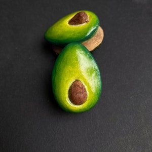 May include: Two avocado-shaped earrings, one green and one brown, with a brown seed in the center. The earrings are made of polymer clay and are about 2 inches long.