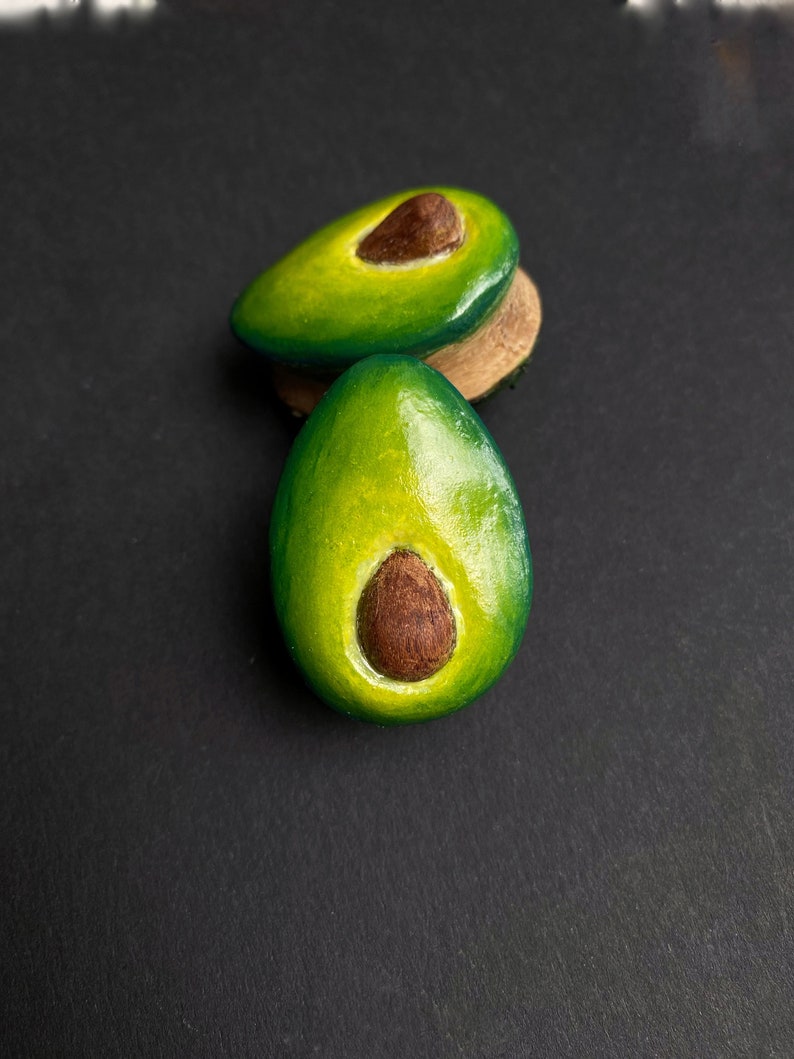 May include: Two avocado-shaped earrings, one green and one brown, with a brown seed in the center. The earrings are made of polymer clay and are about 2 inches long.
