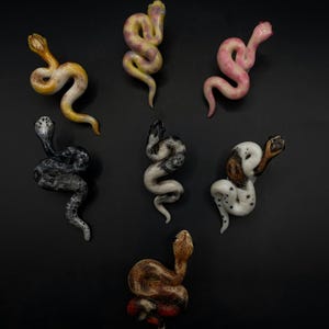 Op de afbeelding: Zeven kleine, handbeschilderde keramische slangfiguren in verschillende kleuren en patronen. De slangen zijn in verschillende posities geposeerd, waaronder opgerold, gekruld en recht.