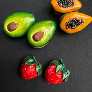 May include: Five miniature clay fruit sculptures: two green avocados, two orange papayas, and one red strawberry. The fruit are arranged on a black surface.