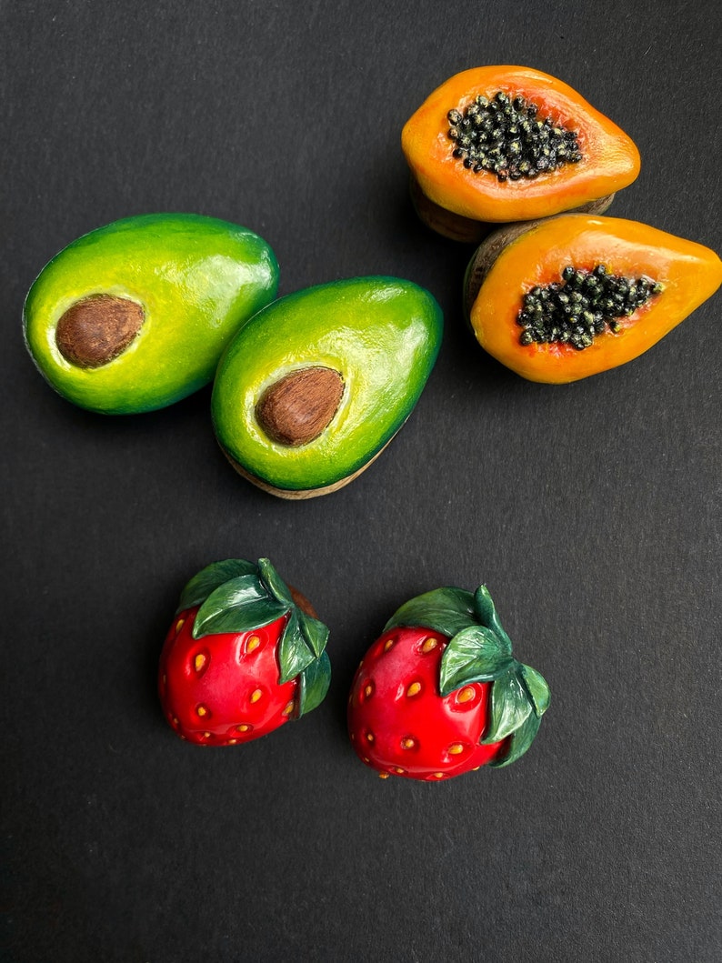 May include: Five miniature clay fruit sculptures: two green avocados, two orange papayas, and one red strawberry. The fruit are arranged on a black surface.
