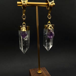 May include: Gold-coloured earrings featuring clear crystal pendants with purple amethyst details. The earrings are displayed on a gold stand with a wooden base, set against a dark background. The design is unique and elegant, suitable for special occasions.