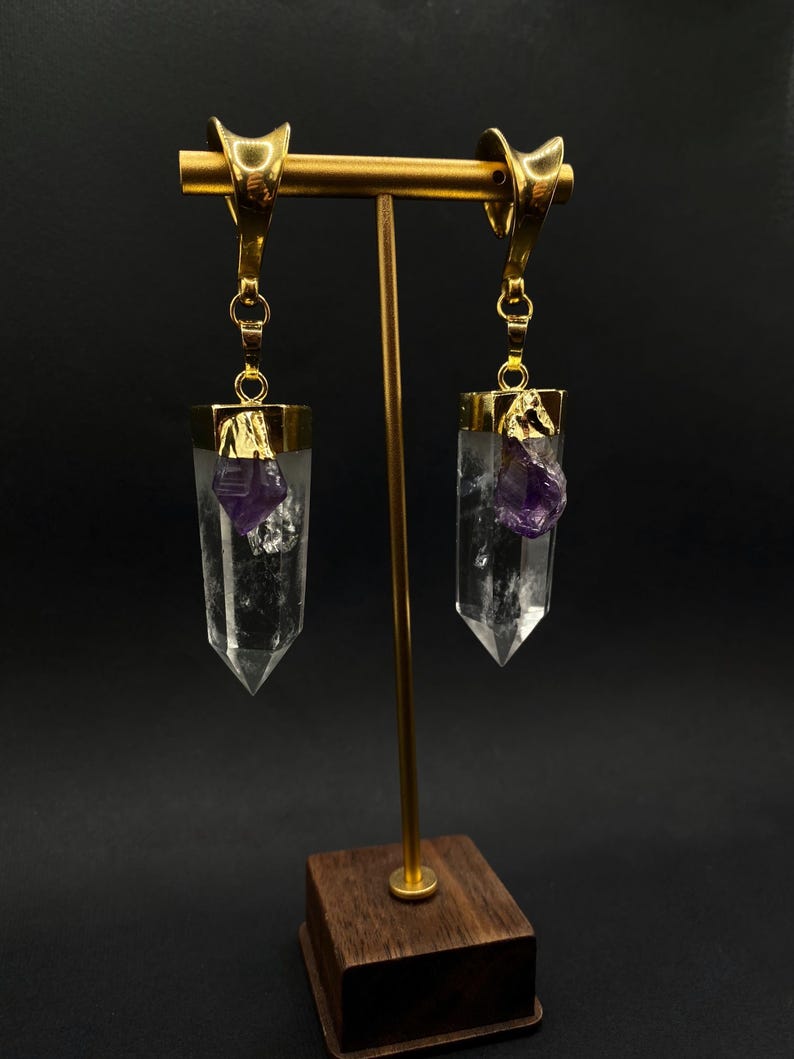 May include: Gold-coloured earrings featuring clear crystal pendants with purple amethyst details. The earrings are displayed on a gold stand with a wooden base, set against a dark background. The design is unique and elegant, suitable for special occasions.