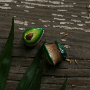 May include: Two avocado-shaped wooden earrings with green and brown paint. The earrings are on a wooden surface with green leaves.