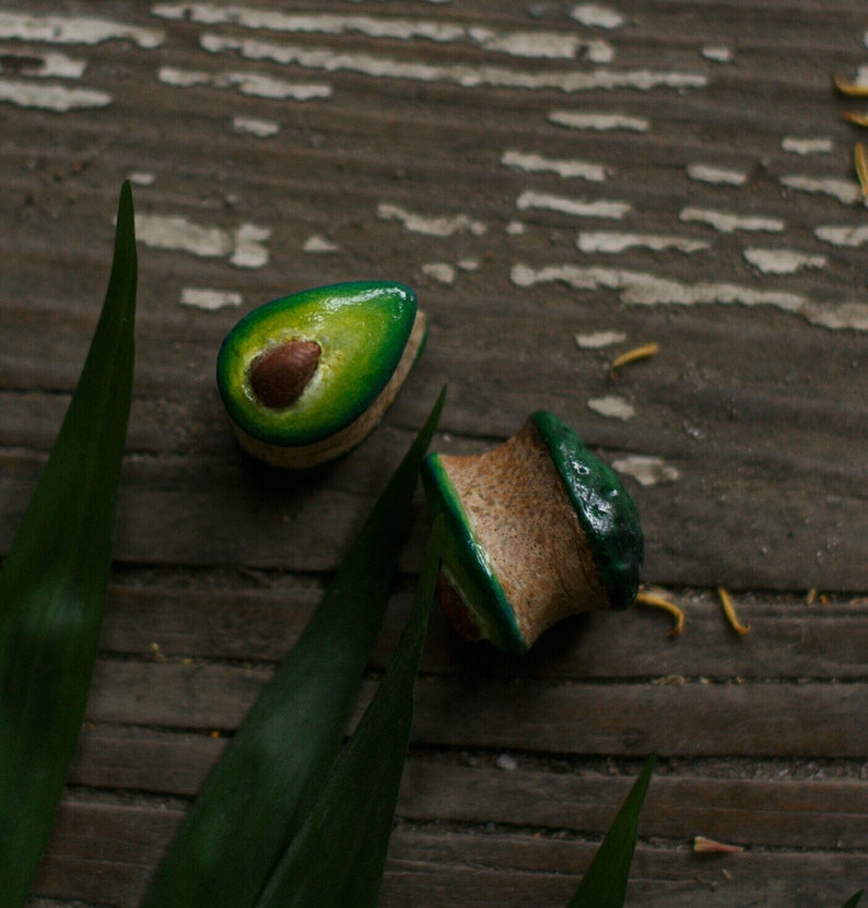 May include: Two avocado-shaped wooden earrings with green and brown paint. The earrings are on a wooden surface with green leaves.